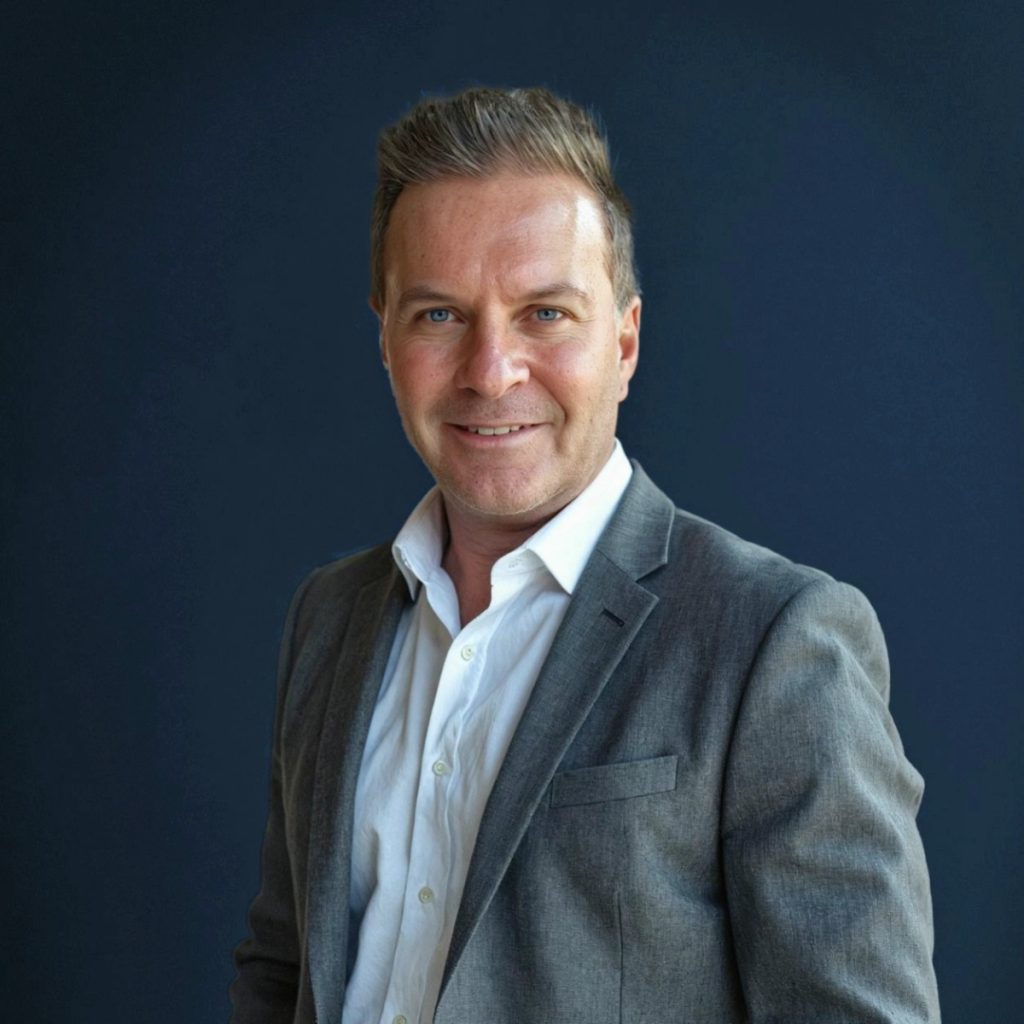 A man in a grey suit jacket and white shirt smiles at the camera, standing against a plain dark blue background.