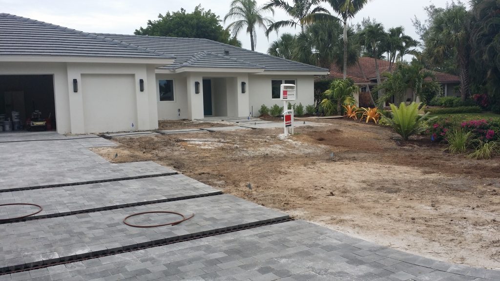 A newly built house with a paved driveway, bare soil yard, and a for-sale sign in front. Neighboring homes and palm trees are visible in the background.
