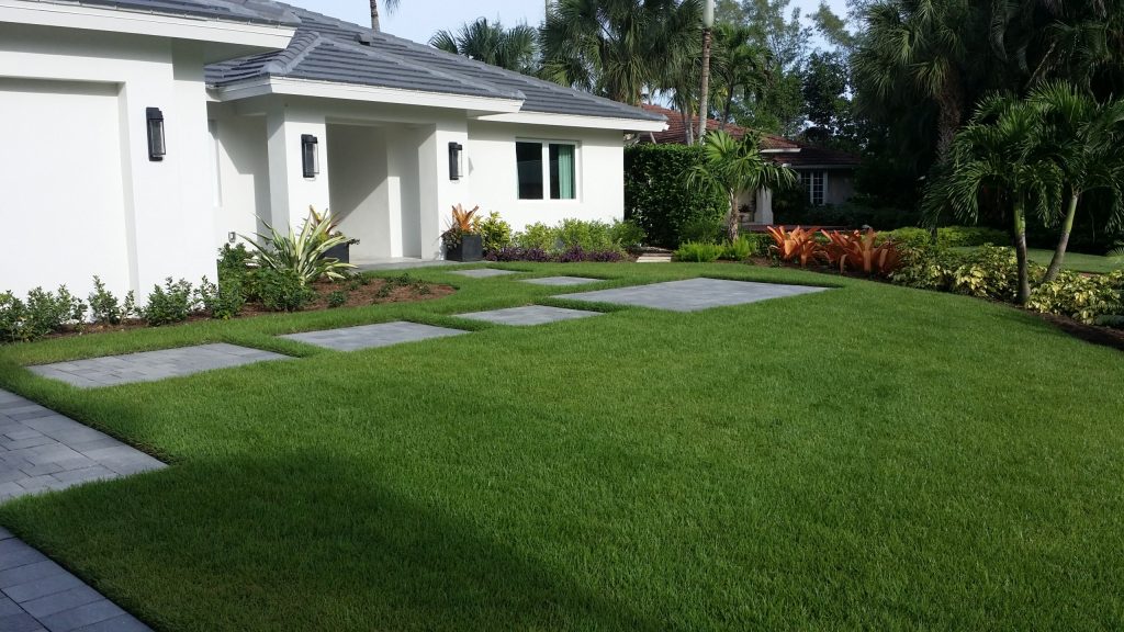 A modern white house with a gray roof, large green lawn, concrete stepping stones, tropical plants, and palm trees in the yard under a sunny sky.