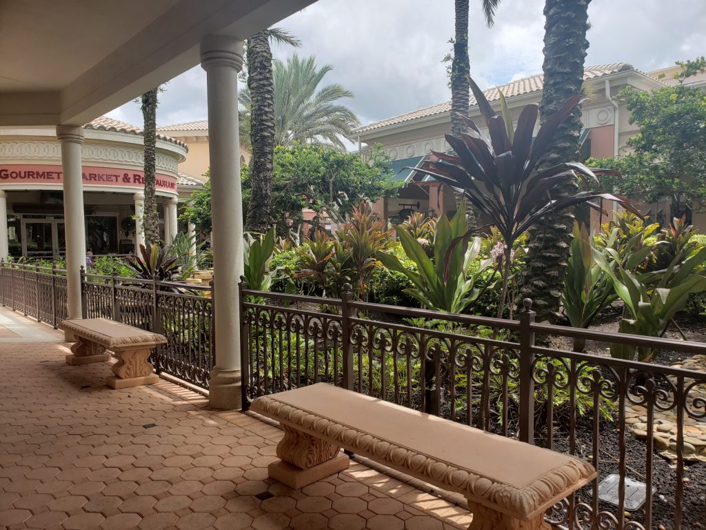 Outdoor walkway with stone benches, decorative iron fence, and lush landscaping featuring palm trees and tropical plants. A building with columns and Gourmet Market & Restaurant sign is visible in the background.