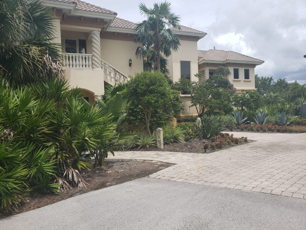 A large beige house with a tiled roof, columns, and balconies is surrounded by palm trees, bushes, and a paved driveway under a cloudy sky.
