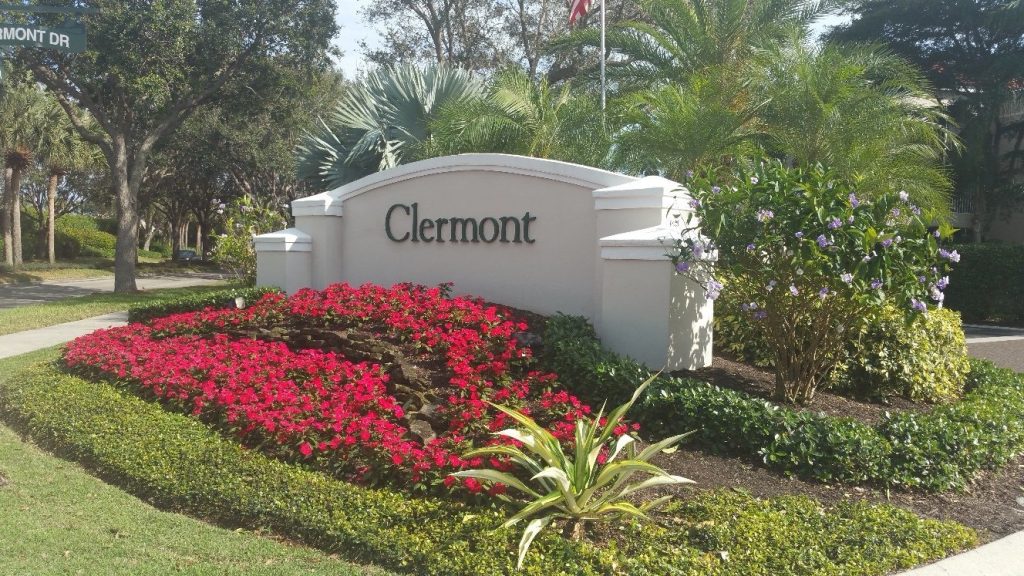 White sign with Clermont written on it, surrounded by neatly trimmed bushes, red flowers, green plants, and palm trees on a sunny day.