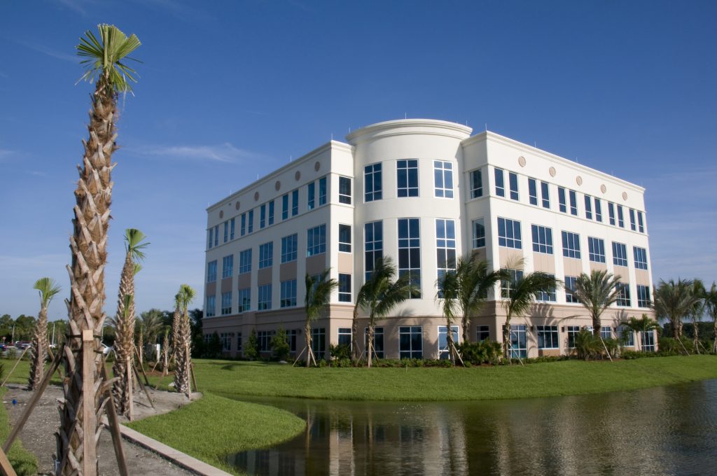 A modern four-story office building with large windows, surrounded by palm trees and a pond, under a clear blue sky.