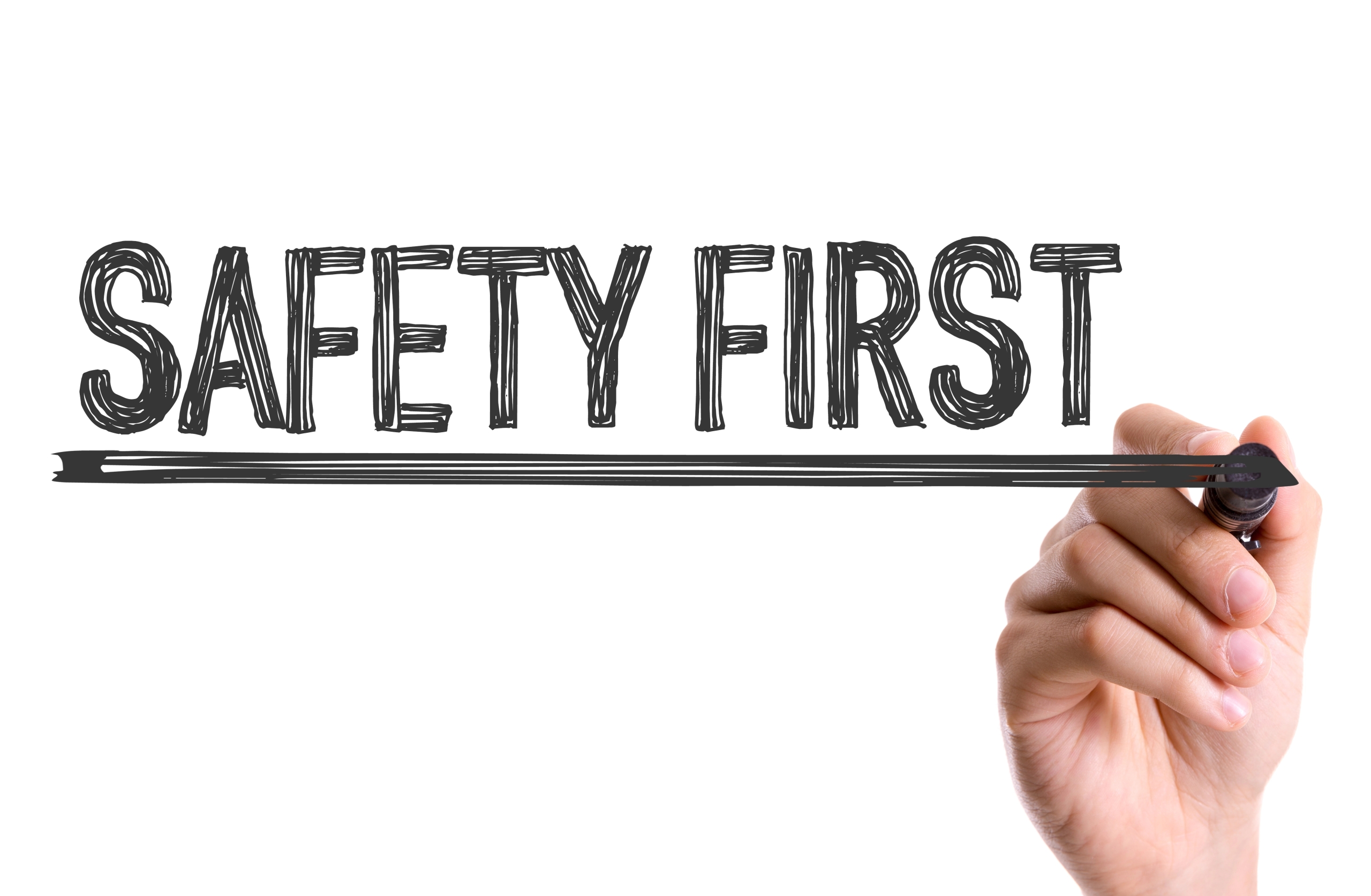 A hand writes SAFETY FIRST in bold, sketch-style letters with a black marker on a transparent surface, underlining the phrase.