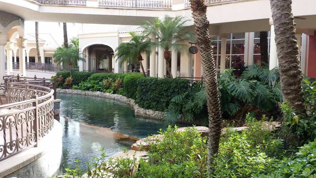 20140407_172049 Scott Whorrall A winding pond bordered by lush plants and palm trees runs alongside a beige building with archways, balconies, and decorative railings.