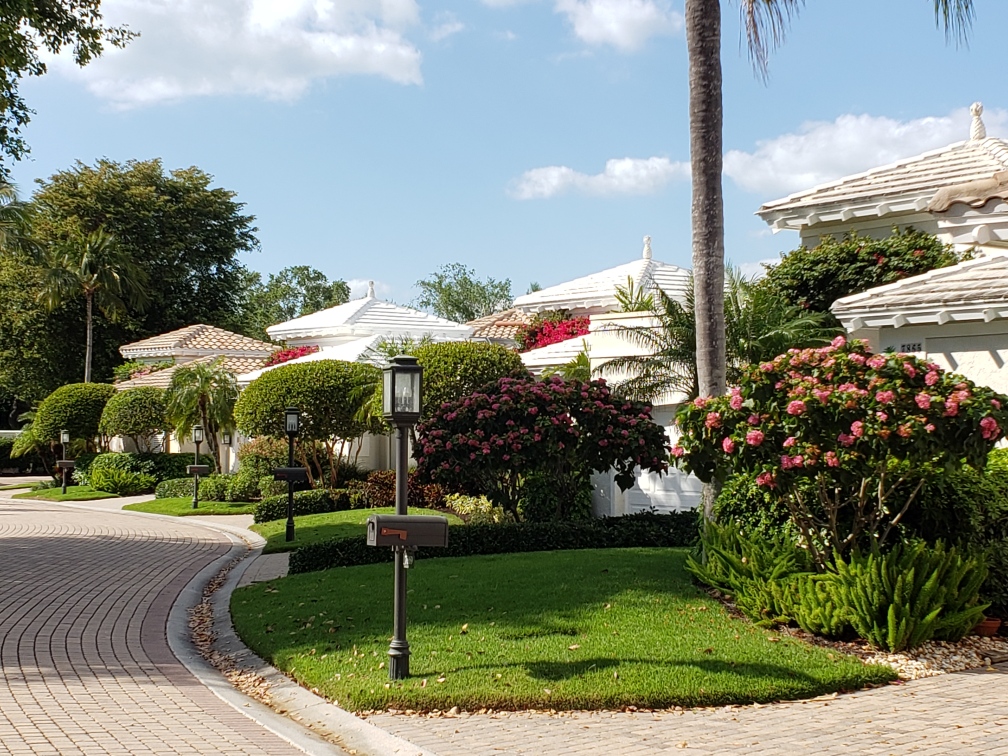 CCB HOA 2 Scott Whorrall A curved street lined with well-manicured lawns, blooming shrubs, palm trees, and white-roofed houses under a partly cloudy sky. A mailbox and streetlight are visible in the foreground.