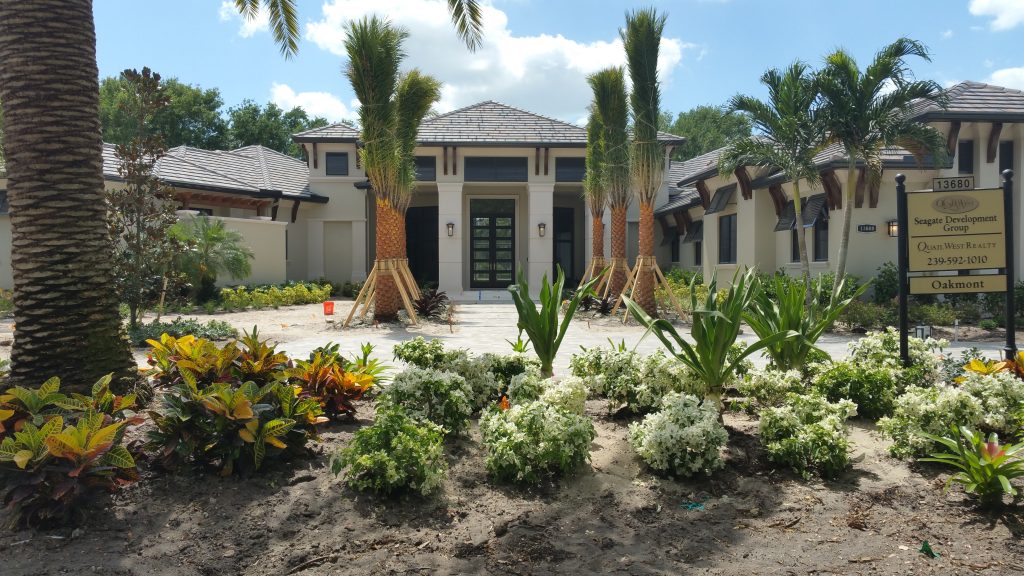 A modern house with a tile roof, surrounded by palm trees and landscaped gardens. A sign in front displays information about the builder and contact details. The sky is partly cloudy.