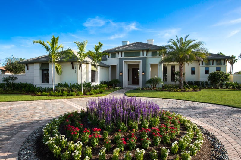 unnamed (7) A modern single-story house with palm trees, a manicured lawn, and a circular flower bed filled with purple, red, and white flowers in the foreground under a clear blue sky.