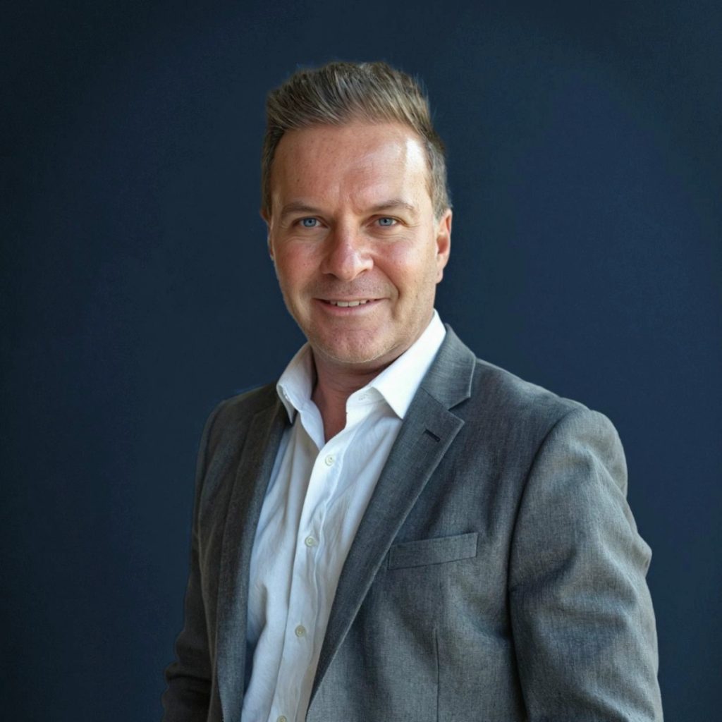 A man wearing a grey suit jacket and white shirt smiles while standing in front of a plain dark blue background.