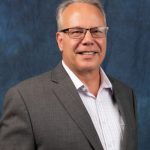Smiling man with short gray hair wearing glasses, a gray suit jacket, and a light plaid shirt, standing in front of a blue textured background.
